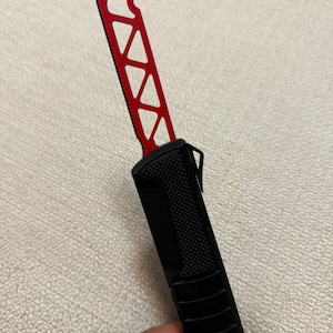 Könnte beinhalten: Ein schwarzes und rotes Messer mit geometrischem Design auf der Klinge. Das Messer hat einen schwarzen Griff mit strukturierter Oberfläche und eine rote Klinge mit einer Reihe von dreieckigen Aussparungen. Die Klinge hat auch einen Flaschenöffner.