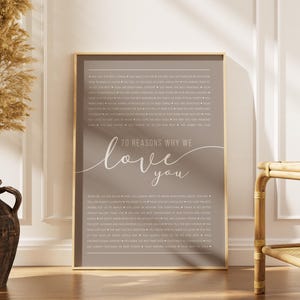 May include: A framed print with a taupe background and gold trim. The print features the text "70 Reasons Why We Love You" in a cursive font. The print is surrounded by a neutral-toned room with a decorative vase and a wooden chair.