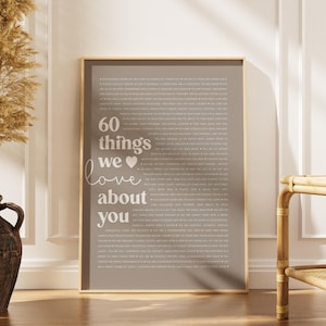 May include: A framed print with a taupe background and gold trim. The print features the text "60 things we love about you" in white script. The background is filled with smaller white text. The print is displayed on a wooden floor.