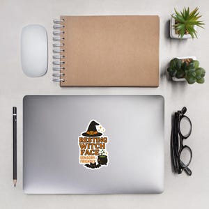 May include: A sticker featuring the text "RESTING WITCH FACE" in orange and white, with a witch hat illustration. Below the text is "SENSORY FRIENDLY" and a cauldron illustration. The sticker is on a laptop.