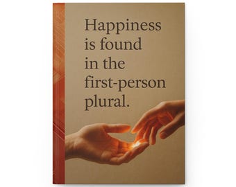 May include: A hardcover journal with the text "Happiness is found in the first-person plural." The cover is a light brown color with a textured spine. The image shows two hands reaching out to each other, with a glowing light between them.
