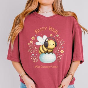 May include: A maroon t-shirt with a graphic of a cartoon bee wearing headphones, sitting on a blue ball. The text "Busy Bee" is above the bee, and "With Sensory Needs" is below. Floral accents surround the bee.