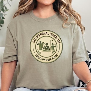 May include: A light olive green t-shirt featuring a circular graphic with the words "Occupational Therapist" and "Function Over Form." The graphic depicts stick figures engaged in various activities. The shirt has short sleeves and a crew neck.