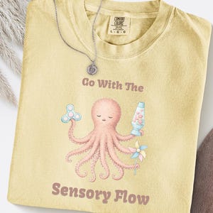 May include: A light yellow Comfort Colors t-shirt with the text "Go With The Sensory Flow" and a cartoon octopus holding a fidget spinner, lava lamp, and pinwheel. A silver necklace with a pendant rests on the shirt.