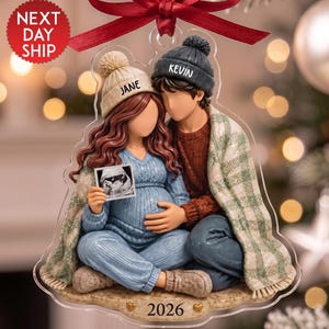 May include: A clear acrylic Christmas ornament featuring a couple sitting together under a green and white plaid blanket. The person on the left wears a beige beanie labeled JANE and a blue sweater, holding a sonogram. The person on the right wears a dark gray beanie labeled KEVIN. The base is inscribed with 2026.