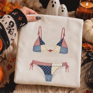 May include: A white t-shirt featuring a graphic print of a two-piece bikini with a patchwork design. The bikini pattern incorporates red and white stripes, blue sections with white stars, and cream-colored panels, connected by a small red star.