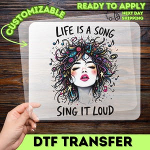 May include: A rectangular DTF transfer sheet featuring a graphic illustration of a person with voluminous, colorful hair intertwined with musical notes and symbols. The text reads LIFE IS A SONG at the top and SING IT LOUD at the bottom in black.