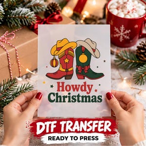 May include: A rectangular DTF transfer sheet featuring a graphic of two cowboy boots, one red and one green, each topped with a cowboy hat. The boots are decorated with ornaments and stars. The text Howdy Christmas is printed in red and green.