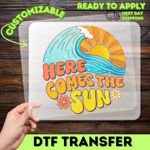 May include: A clear DTF transfer sheet featuring a graphic design of a large ocean wave and a bright yellow sun with orange rays. The text reads HERE COMES THE SUN in bold, rounded orange and yellow letters, accented by two small pink flowers.
