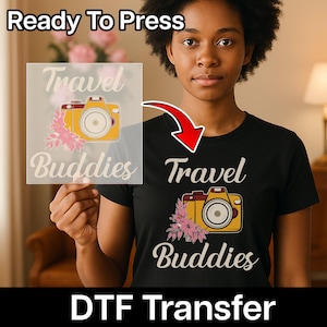 May include: A black t-shirt with a yellow camera graphic and the words "Travel Buddies". The image also shows a "Ready To Press" DTF transfer with the same design. The transfer is held up to the shirt, with a red arrow pointing to the shirt.