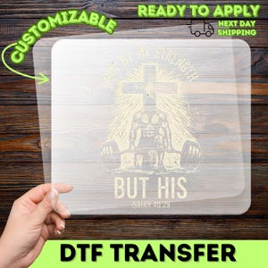 May include: A DTF transfer with a religious theme. The design features a weightlifter, a cross, and the text "NOT BY MY STRENGTH BUT HIS ISAIAH 40:29". The transfer is customizable and ready to apply.
