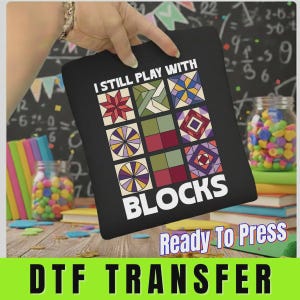 May include: A black fabric transfer sheet featuring the white text I STILL PLAY WITH BLOCKS above and below a grid of nine colorful geometric quilt-style square patterns in shades of red, green, purple, yellow, and blue on a dark background.