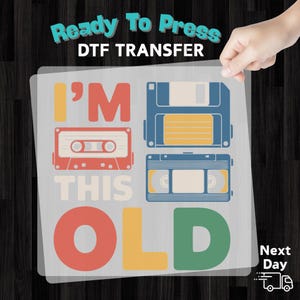 May include: A DTF transfer featuring the phrase "I'M THIS OLD" with retro graphics of a cassette tape, floppy disk, and VHS tape in red, blue, and yellow. The words "Ready To Press" are at the top.