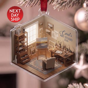 May include: A clear, hexagonal acrylic ornament featuring a detailed miniature art studio scene. The interior displays a wooden easel with a landscape painting, a desk with brushes and paints, a bookshelf, and a chair. Text reads Carol's ART STUDIO 2026.