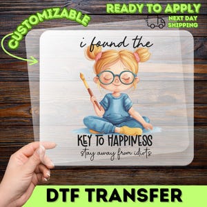 May include: A clear DTF transfer sheet featuring a cartoon illustration of a blonde person with glasses and hair buns, holding a paintbrush. The text reads: i found the KEY TO HAPPINESS stay away from idiots. The design is printed on a transparent film.