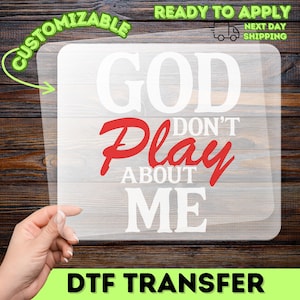 May include: A clear DTF transfer with the text "GOD DON'T PLAY ABOUT ME" in white and red. The word "CUSTOMIZABLE" is in green, and "READY TO APPLY" is in green with a truck icon. The transfer is held by a hand against a wood grain background.