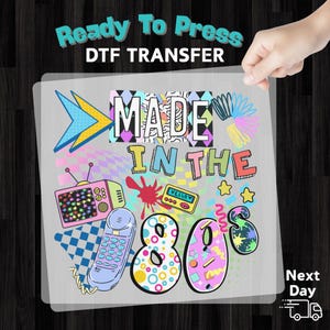 May include: A DTF transfer sheet with a colorful 80s theme. The design includes the text "MADE IN THE 80s" with a retro television, telephone, and cassette tape. The sheet is held by a hand, with the words "Ready To Press" at the top.