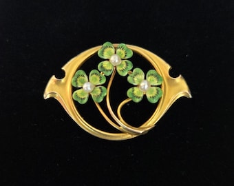 May include: Gold-toned brooch featuring three green and white enameled shamrock flowers with pearl centers. The shamrocks are set within a gold oval frame. A vintage-style jewelry piece.