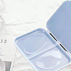 May include: An open, light blue, hinged case with two clear, oval-shaped compartments. The case is on a white, textured surface. The case is empty.