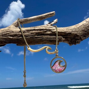 Pode incluir: Um colar dourado com um design de onda. O colar apresenta um pingente com formato de onda preenchido com fragmentos rosa e branco. Está pendurado em um pedaço de madeira flutuante contra um céu azul.