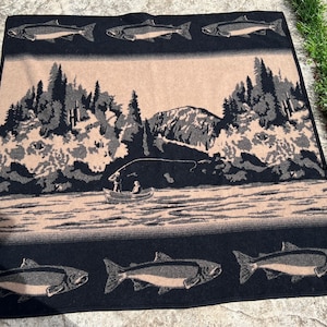 Puede incluir: Una manta tejida rectangular con un borde negro y motivos repetidos de peces. El panel central muestra un paisaje en tonos beige y negro con un bosque, monta&ntilde;as y una peque&ntilde;a barca con dos figuras sobre un r&iacute;o, enmarcado por peces.