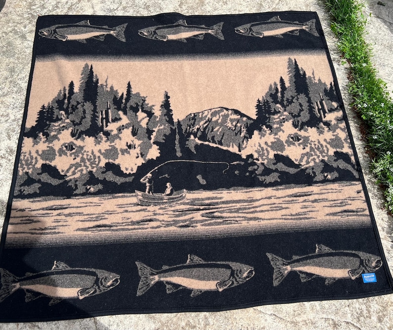 Puede incluir: Una manta tejida rectangular con un borde negro y motivos repetidos de peces. El panel central muestra un paisaje en tonos beige y negro con un bosque, monta&ntilde;as y una peque&ntilde;a barca con dos figuras sobre un r&iacute;o, enmarcado por peces.