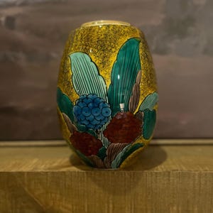 Puede incluir: Un jarrón decorativo de cerámica con fondo dorado y diseño floral. El jarrón presenta flores y hojas turquesas, azules y granates. El jarrón mide aproximadamente 15 cm de altura.