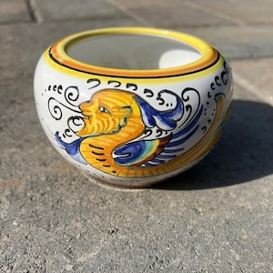 Puede incluir: Un pequeño cuenco redondo de cerámica con base blanca, adornado con un diseño de criatura mítica amarilla y azul. El borde está delineado en amarillo y negro. El cuenco probablemente se utiliza con fines decorativos.