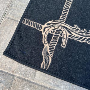 Op de afbeelding: Een zwart rechthoekig tapijt met een beige grafisch ontwerp van een touw dat om een hoefijzer is geknoopt. Het tapijt heeft een afgewerkte zwarte stoffen rand langs de zijkanten en een getextureerd oppervlak. Het ontwerp staat centraal op de donkere achtergrond.
