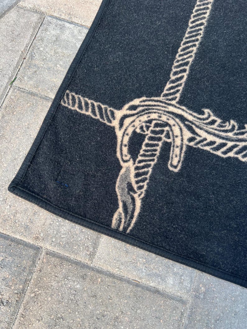 Op de afbeelding: Een zwart rechthoekig tapijt met een beige grafisch ontwerp van een touw dat om een hoefijzer is geknoopt. Het tapijt heeft een afgewerkte zwarte stoffen rand langs de zijkanten en een getextureerd oppervlak. Het ontwerp staat centraal op de donkere achtergrond.