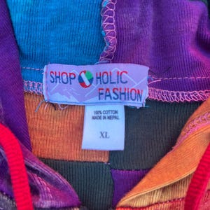 K&ouml;nnte beinhalten: Ein Patchwork-Kapuzenpullover mit Rei&szlig;verschluss in einer lebendigen Farbpalette aus Lila, Orange, Blau und Dunkelgr&uuml;n. Ein wei&szlig;es Etikett zeigt den Text SHOP HOLIC FASHION, 100% COTTON, MADE IN NEPAL und XL. Das Kleidungsst&uuml;ck hat rote Kordelz&uuml;ge.
