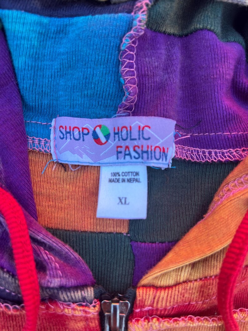 K&ouml;nnte beinhalten: Ein Patchwork-Kapuzenpullover mit Rei&szlig;verschluss in einer lebendigen Farbpalette aus Lila, Orange, Blau und Dunkelgr&uuml;n. Ein wei&szlig;es Etikett zeigt den Text SHOP HOLIC FASHION, 100% COTTON, MADE IN NEPAL und XL. Das Kleidungsst&uuml;ck hat rote Kordelz&uuml;ge.