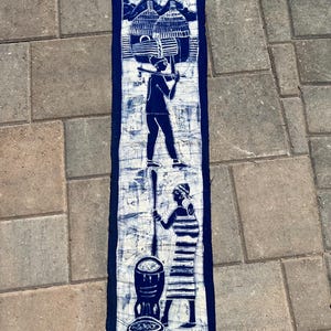 May include: A rectangular fabric wall hanging featuring a dark blue and white batik print. The design depicts two stacked scenes: a figure carrying a large drum on their head above a figure standing beside a drum and a patterned bowl.