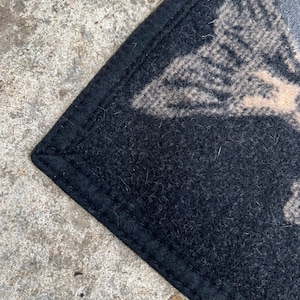 Puede incluir: Una alfombrilla de fieltro negro con un borde cosido. La superficie presenta un dise&ntilde;o gr&aacute;fico parcial en color beige claro o bronceado, mostrando un patr&oacute;n texturizado que se asemeja a una forma de ala o pluma sobre el fondo negro oscuro.