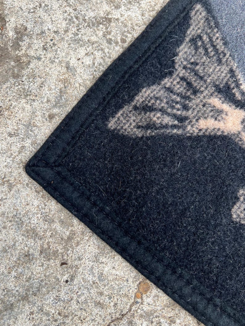 Puede incluir: Una alfombrilla de fieltro negro con un borde cosido. La superficie presenta un dise&ntilde;o gr&aacute;fico parcial en color beige claro o bronceado, mostrando un patr&oacute;n texturizado que se asemeja a una forma de ala o pluma sobre el fondo negro oscuro.