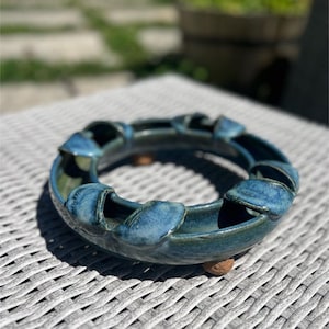 Puede incluir: Cenicero circular de cerámica hecho a mano en tonos azules y verdes. El cenicero presenta un diseño único con elementos elevados en forma de pétalos alrededor del borde. Descansa sobre pequeños pies redondeados.
