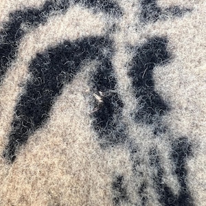 Op de afbeelding: Een close-up van een textieloppervlak met een pluizige textuur met een hoge pool. Het materiaal vertoont een contrasterend patroon van diepzwarte, onregelmatige, organische vormen tegen een achtergrond van lichtbeige, vezelig materiaal.