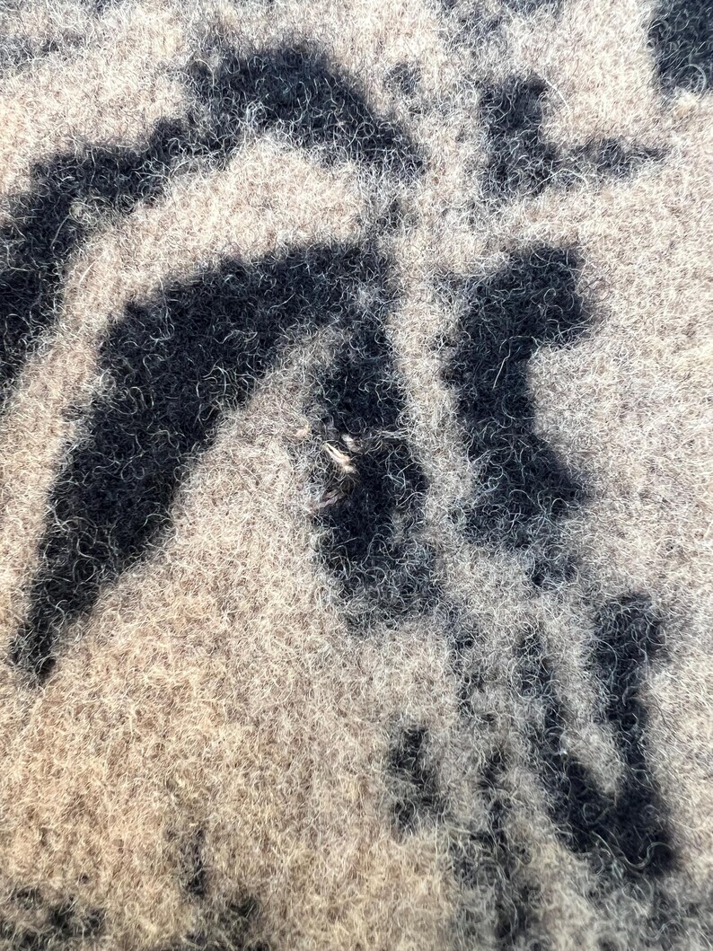 Op de afbeelding: Een close-up van een textieloppervlak met een pluizige textuur met een hoge pool. Het materiaal vertoont een contrasterend patroon van diepzwarte, onregelmatige, organische vormen tegen een achtergrond van lichtbeige, vezelig materiaal.