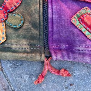 K&ouml;nnte beinhalten: Eine Jacke mit Rei&szlig;verschluss im Patchwork-Design mit Stoffbahnen in Olivgr&uuml;n und Violett. Das Kleidungsst&uuml;ck verf&uuml;gt &uuml;ber dekorative Stoffapplikationen mit sichtbaren blauen und gr&uuml;nen N&auml;hten, einen schwarzen Rei&szlig;verschluss und zwei geknotete orangefarbene Kordeln am Saum.