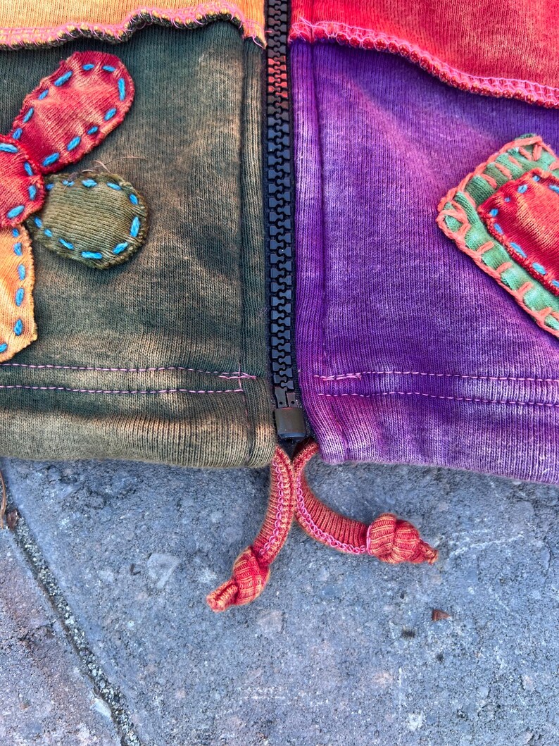 K&ouml;nnte beinhalten: Eine Jacke mit Rei&szlig;verschluss im Patchwork-Design mit Stoffbahnen in Olivgr&uuml;n und Violett. Das Kleidungsst&uuml;ck verf&uuml;gt &uuml;ber dekorative Stoffapplikationen mit sichtbaren blauen und gr&uuml;nen N&auml;hten, einen schwarzen Rei&szlig;verschluss und zwei geknotete orangefarbene Kordeln am Saum.