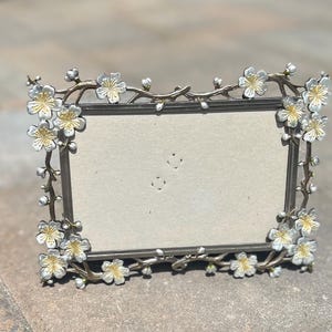 May include: A rectangular photo frame with a floral design. The frame is silver-toned with delicate branches and white and gold flower accents. The frame has a neutral-colored backing and is designed to hold a photograph.