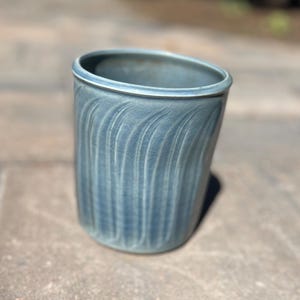 Peut inclure: Un gobelet en céramique bleu avec un motif de vagues verticales texturées. Le bord est lisse et de couleur plus claire. La tasse faite à la main mesure environ 10 cm de haut.