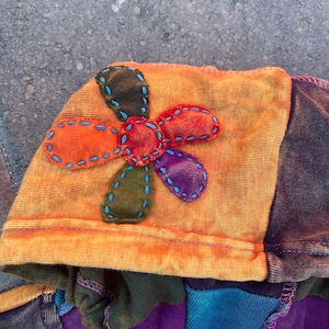 K&ouml;nnte beinhalten: Ein Kleidungsst&uuml;ck mit Kapuze aus orange-gelbem Batikstoff mit einer mehrfarbigen Stoffblumenapplikation. Die Blume hat Bl&uuml;tenbl&auml;tter in Orange, Rot, Violett und Olivgr&uuml;n, die mit blauen N&auml;hten umrandet sind. Ein Etikett tr&auml;gt die Aufschrift SHOP HOLIC.