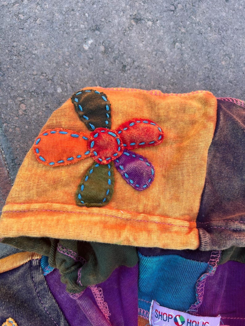 K&ouml;nnte beinhalten: Ein Kleidungsst&uuml;ck mit Kapuze aus orange-gelbem Batikstoff mit einer mehrfarbigen Stoffblumenapplikation. Die Blume hat Bl&uuml;tenbl&auml;tter in Orange, Rot, Violett und Olivgr&uuml;n, die mit blauen N&auml;hten umrandet sind. Ein Etikett tr&auml;gt die Aufschrift SHOP HOLIC.