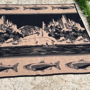 Puede incluir: Una manta tejida rectangular con un dise&ntilde;o en negro y beige. El patr&oacute;n muestra un paisaje central de una persona en un bote pescando en un lago rodeado de &aacute;rboles y monta&ntilde;as, bordeado por filas de peces en los bordes superior e inferior.