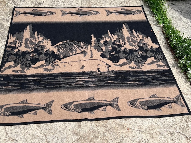 Puede incluir: Una manta tejida rectangular con un dise&ntilde;o en negro y beige. El patr&oacute;n muestra un paisaje central de una persona en un bote pescando en un lago rodeado de &aacute;rboles y monta&ntilde;as, bordeado por filas de peces en los bordes superior e inferior.