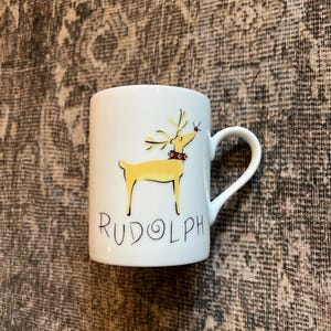 以下が含まれることがあります： 黄色いトナカイのイラスト、赤い首輪と鼻が描かれた白いセラミックマグ。 「RUDOLPH」の文字が黒い筆記体で書かれています。 マグカップにはカーブしたハンドルが付いています。 クリスマスをテーマにしたアイテム。