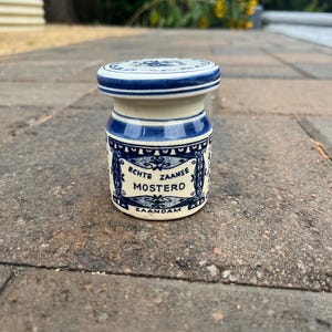 Puede incluir: Un pequeño tarro de mostaza de cerámica color crema con tapa a juego, decorado con patrones azules y el texto ECHTE ZAANSE MOSTERD ZAANDAM. El tarro tiene un cuerpo cilíndrico con un borde ligeramente más ancho y una tapa plana circular.