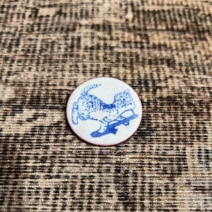 Puede incluir: Un botón redondo blanco con una ilustración azul de un gato en monopatín. El botón tiene un borde rojo. El gato se representa en una pose juguetona. El fondo es una tela texturizada de tonos neutros.