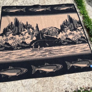 Puede incluir: Una manta tejida rectangular con un esquema de color negro y bronceado. El diseño muestra un paisaje central de un lago, un pequeño bote con dos figuras y montañas. Los bordes superior e inferior presentan un patrón repetitivo de peces.